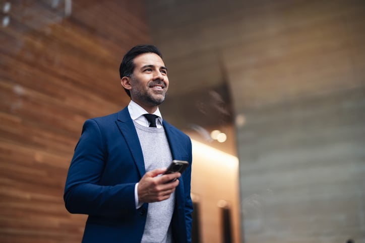 Confident Businessman In Blue Suit Smiling While Holding Smartphone In A Modern Indoor Lobby